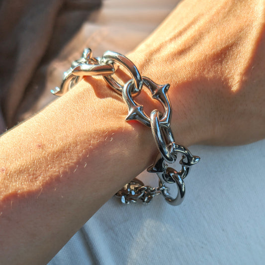 A close-up of a person's wrist wearing a chunky stainless steel chain bracelet with star-shaped links, lit by warm sunlight against a white shirt background.