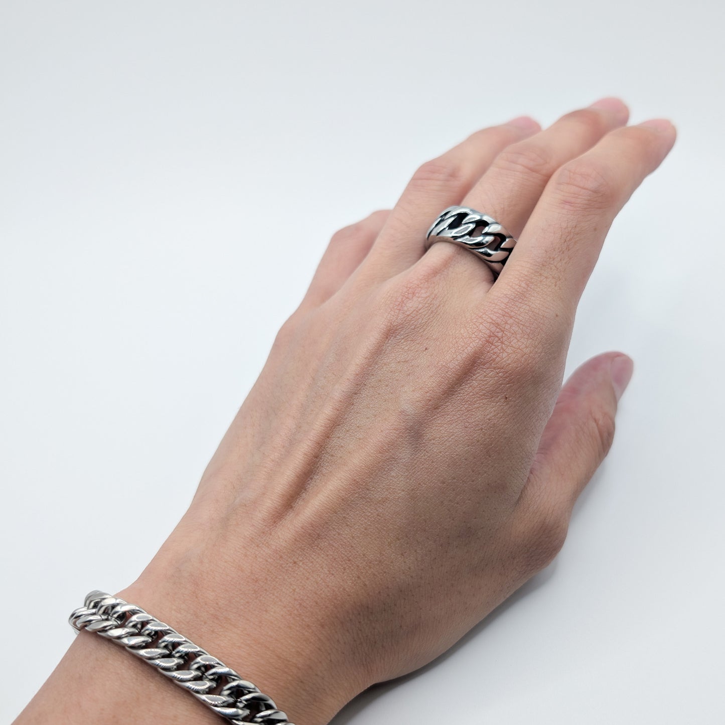 A close-up of a person's hand wearing a chunky silver-toned curb link ring and a matching silver-toned curb link bracelet against a plain light gray background.