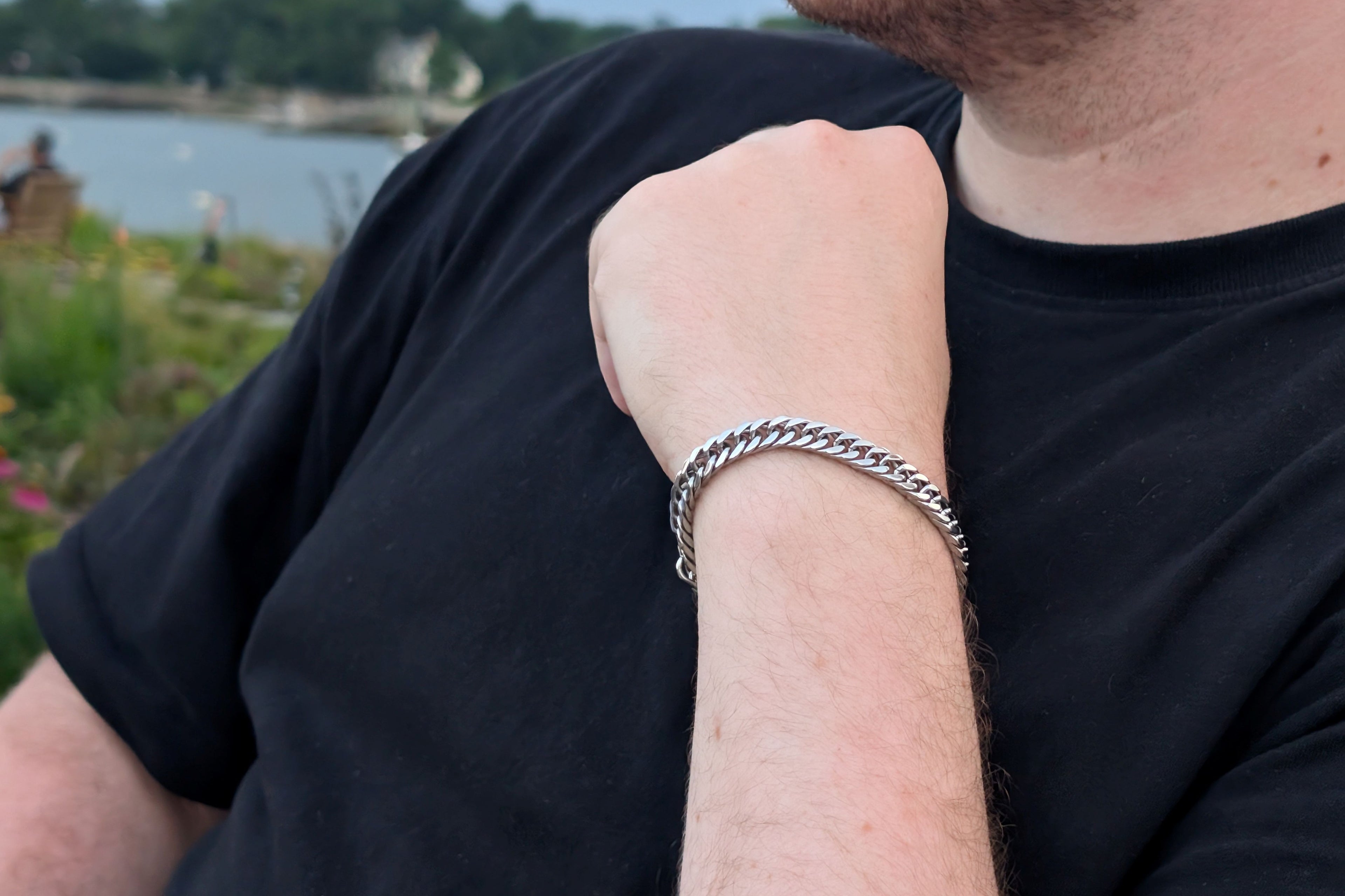 A close-up of a man's forearm wearing a silver-toned cuban link chain bracelet, wearing a black t-shirt with an outdoor waterfront background.